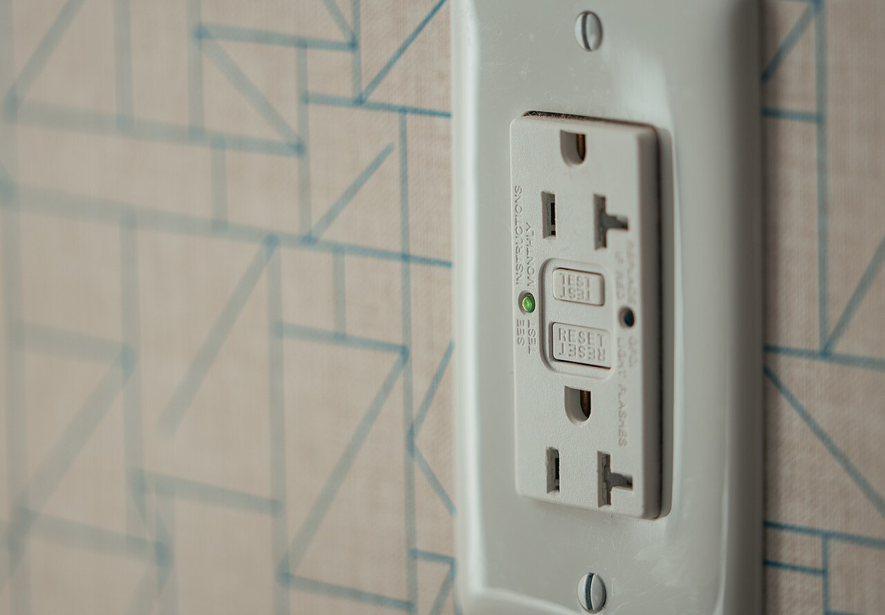 A white GFCI electrical outlet with test and reset buttons and a green indicator light, mounted in a standard electrical box