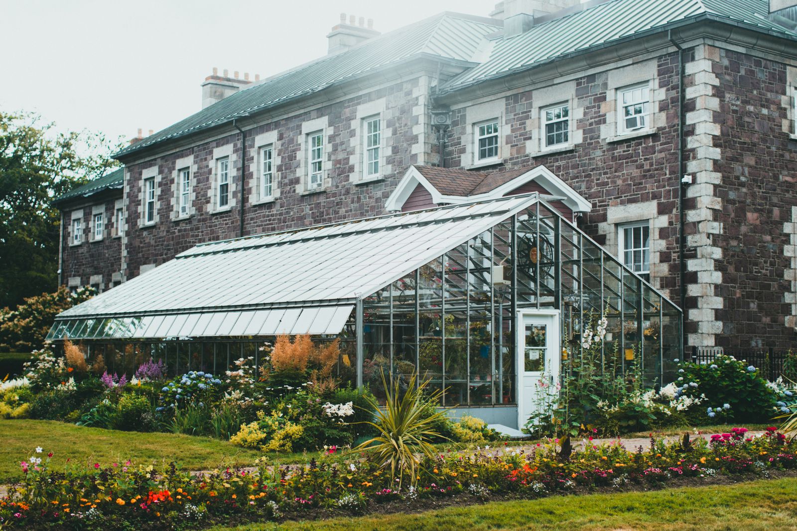 A picturesque greenhouse surrounded by vibrant flowering gardens against a historic stone building in a heritage garden setting