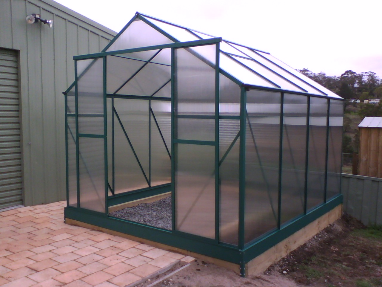A small polycarbonate-covered metal greenhouse structure showing the rigid twin-wall panel construction and aluminum framing